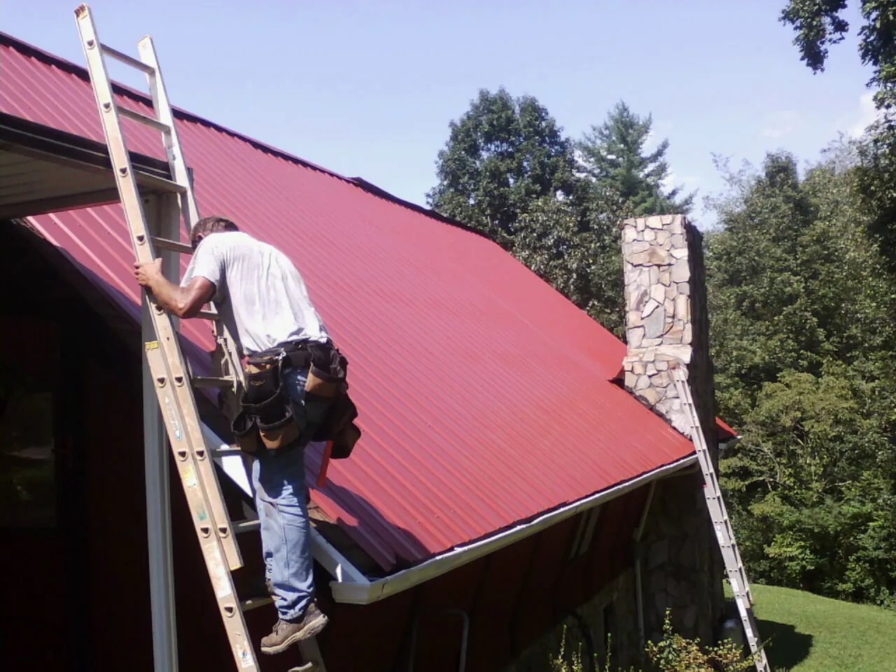 Roofer on a ladder beside a red metal roof and stone chimney in Taylor